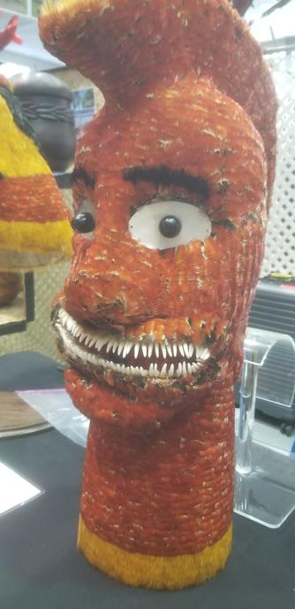 A table with some items made from feathers. One looks like a giant head with bulbous eyes it is made from small, smooth, red feathers. The face has a mouth full of shark's teeth.