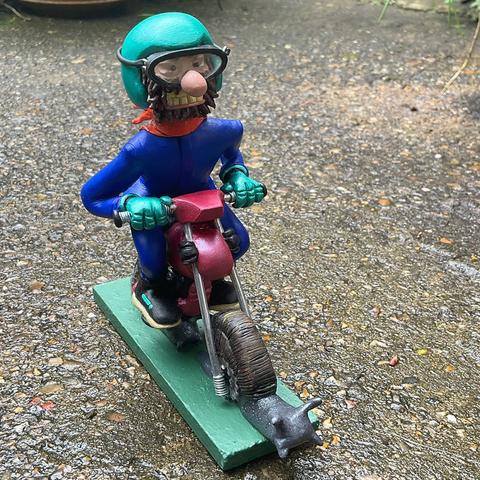 A grimacing biker wearing goggles rides a motorbike with 2 Snails instead of wheels. He has a shiny turquoise helmet a scruffy beard and orange scarf.