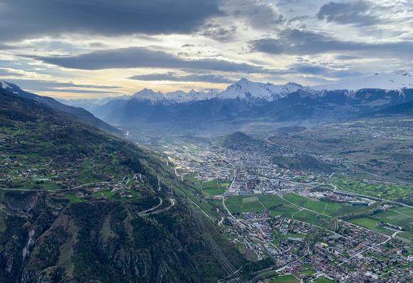 View over a wide valley in which an Alpine town lies and which is lined with high mountains whose peaks are still covered in snow.
