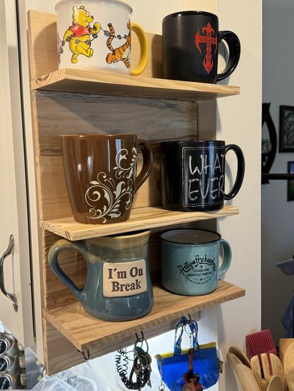 A collection of various mugs with different designs and inscriptions displayed on wooden shelving, with some hanging keys visible at the bottom.