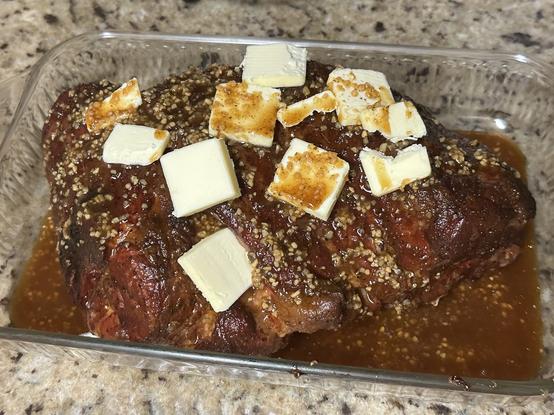 A seasoned pork butt in a glass dish topped with butter and garlic and a marinade sauce
