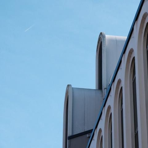 The arches of a modern building’s roofline contrast against a clear blue sky with a faint airplane trail.