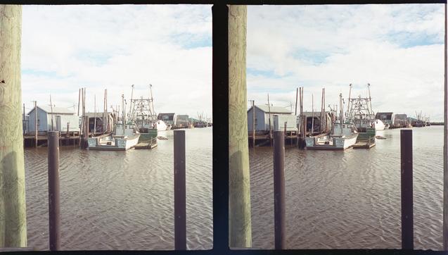 3D photo (two frames side-by-side) of a couple of fishing boats docked in harbor, with a wooden piling and two metal posts standing in the foreground.