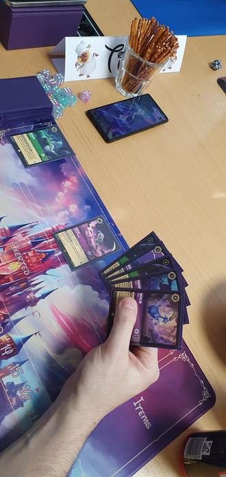 Ein Mitspielender hält einige Lorcana-Karten in der Hand. Unter der Hand ist eine Spielmatte. Einige Karten liegen darauf. Im Hintergrund sieht man den Deck-Stapel, ein paar Würfel und ein paar Snacks.