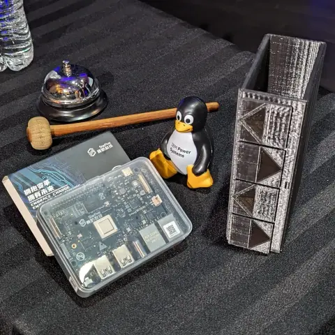Table with VisionFive 2 board, IBM Power squishy tux penguin, 3d-printed IBM z16 and an unrelated bell.