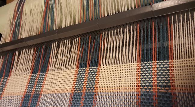 Un tissu en cours de tissage sur un métier à peigne envergeur.  Sur une chaine de ton blanc / crème,  une succession de lignes bleu pétrole foncé et vieux rose foncé vont créer des carreaux en rencontrant une trame crème / bleu clair / vieux rose pâle