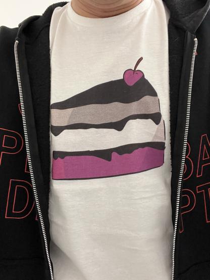 A person wearing a white t-shirt with a graphic of a slice of cake and a cherry on top, partially covered by a black zip-up hoodie. The cake is in the colors of the Asexual flag.