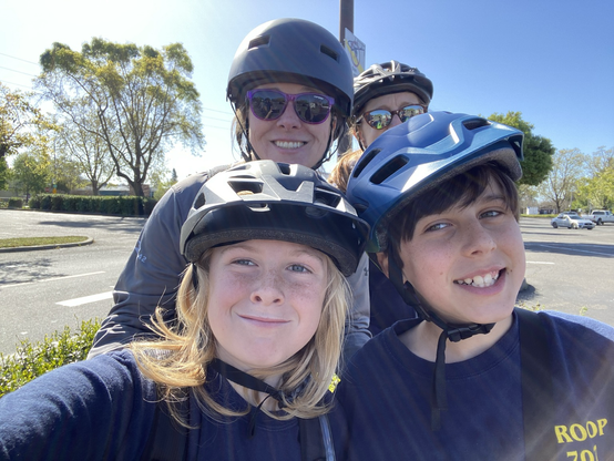 Two kids smiling with two ladies smiling in background all with bike helmets on.