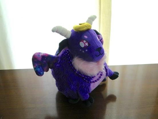 A purple goat plushie -- with cosmic wings, a pink beard, with a gold Moon halo and white horns -- and wearing a purple beaded necklace.