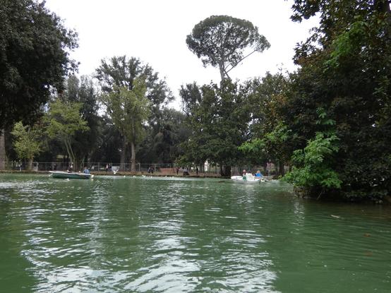 Bounded by tall trees, a small boating lake reflects the light. One or two boats dot the surface.