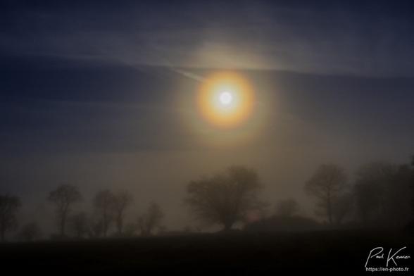 Photo, en couleur, de nuit, dans une brume très épaisse, au-dessus de la campagne, dont on devine quelques arbres en bas de la photo. Au-dessus des arts, on devient également la pleine lune, à travers la prune, qui génère une magnifique couronne autour de notre satellite, couronne très colorée avec les couleurs de l'arc-en-ciel en cercles concentriques.