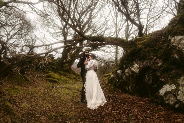 In the image, a couple stands intimately embraced in a serene, natural landscape. It appears to be a cool or misty day. They are in a forest setting, surrounded by trees with gnarled, bare branches suggesting late autumn or winter. The trees are covered in green moss, adding a touch of vibrant color to the otherwise muted tones of the scene. The ground is carpeted with fallen leaves, and there are rocks covered with moss, indicating a moist environment, perhaps near the coast or in a hilly area. The man, with a full beard, wears dark attire, providing a stark contrast to the woman's white, vintage-style dress. The mood is moody and romantic, with a timeless feel, as they stand in an intimate pose, isolated from the bustle of the world. The image encapsulates a moment of tranquility and connection between two people in the quiet of nature.