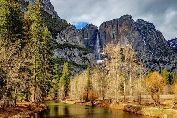 Merced River and Yosemite Falls

This is an image of Yosemite Falls and the Merced River taken from the swinging bridge in Yosemite Valley.
The foreground consist of the Merced River. The river is relatively tranquil as it is winter and the water from the falls is reduced due to the cold temperatures at higher elevation.
On the left bank of the river grows tall pine trees, mostly Douglas Firs. On the right side of the river the banks are lined with hardwood trees. These trees have lost their leaves for the winter.
At the base of the upper falls you can see an ice cone that has built up during the winter. The shear rock cliffs frame the falls. 
The sky is full of big fluffy clouds with just a hint of blue sky just above the falls.