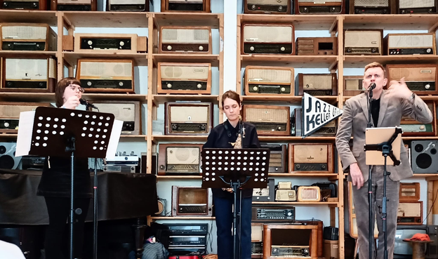 Im Vordergrund die Musiker, links die Sängerin, in der Mitte die Saxophonistin und rechts der Sänger.
Im Hintergrund ein hohes Regal mit ganz vielen alten Radioapparaten.