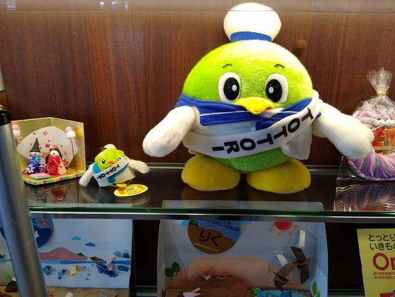 鳥取の梨のマスコットのぬいぐるみ（大小）。ガラスの向こう側にある。セーラー服に水兵帽、胸には「TOTTORI」の帯。