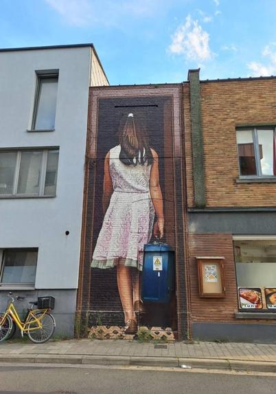 Streetartwall. An oversized mural of a young woman with a suitcase was sprayed/painted on a single-storey brick wall between two buildings. She can be seen from behind. She has long brown hair, wears a short summer dress with a pink and white pattern and holds a suitcase in her hand. She seems to be going into the house. No one would recognize that the suitcase is a transformer station if it weren't for a warning sticker with a lightning bolt. 
Info: The artwork is part of the "Tour Elentrik". This is a nationwide project in which old and ugly electricity and utility boxes are redesigned by local creatives and professional artists from the Treepack network.