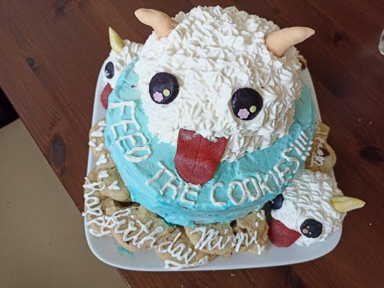 A blue cake, where on top there is a big poro (cute, fluffy, ball like animal from league of legends). Next to the poro there is a text saying "feed the cookies!!!!".
On the plate around the cake are cookies and two more smaller poros. On the cookies there is a text saying "happy birthday Mimi"