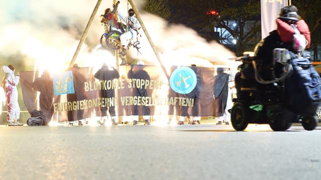 2 Menschen hängen in einem Dreibein , eine der 2 Personen im Rolltuhl. es ist Nacht. die Demonstrierenden tragen weiße Anzüge und haben Rauchtöpfe angezündet, der Rauch davon ist im Bild. auf dem Banner vor dem Tripod steht Blutkohle stoppen Energiekonzerne vergesellschaften und das Logo von Ende Gelände. im Vordergrund eine Peron im E-Rollstuhl. Das Bild wurde mit Langzeitbelichtung aufgenommen.