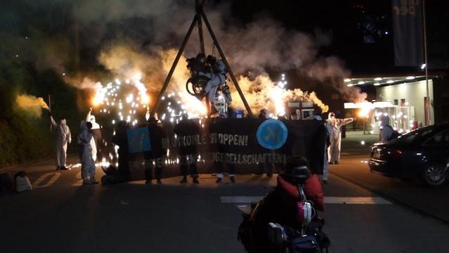 2 Menschen hängen in einem Dreibein , eine der 2 Personen im Rolltuhl. es ist Nacht. die Demonstrierenden tragen weiße Anzüge. auf dem Banner vor dem Tripod steht Blutkohle stoppen Energiekonzerne vergesellschaften und das Logo von Ende Gelände. Bild in Langzeitbelichtung aufgenommen, im Bild auch der Rauch von bunten Rauchtöpfen.