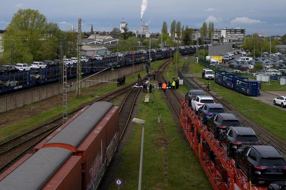 Das Bild wird dominiert von Zügen, ein langer Zug voller PKW erstreckt sich von der linken Bildkante im Hintergrund bis zum Mercedes-Werk am Horizont. Vorne links und rechts ragen zwei weitere Züge ins Bild. Rechts sind PKW auf einem Parkplatz zu sehen. In der Mitte auf den Gleisen sind mehrere Menschen, teilweise in Warnwesten zu sehen. Dazwischen liegen Transparente. Zwischen den Gleisen wächst grünes Gras.
Bild: Pay Numrich
