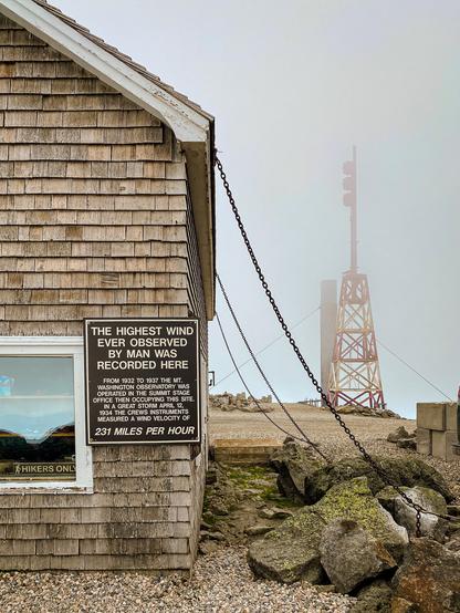 Color photo of a wood building being held down  by 3 heavy chains on the summit of a very windy Mt Washington.  Windy and cloudy so the antenna tower behind the building not that clear. 

The sign on the building reads:
The Highest Wind ever observed by man was recorded here. 

From 1932 to 1937 the Mt Washington Observatory was operated in teh Summit Stage office then occupying this site. In a Great Storm April 12, 1934 the crews instruments measured a wind velocity of
231 MILES PER HOUR
