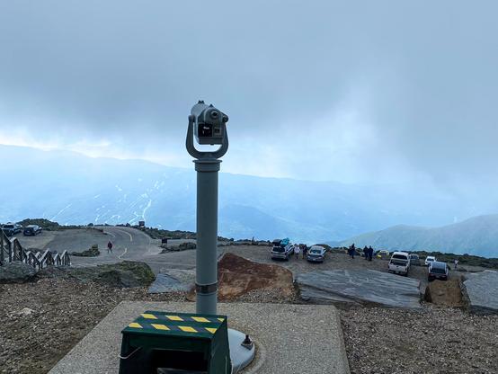 A color photo from the summit looking out over the parking lot and other mountains in the range.  You can see the lower edge of the clouds we were in moving past.