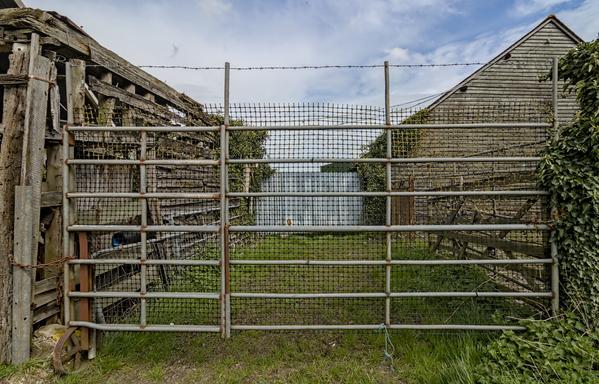A photograph of a metalic farm gate. There are wooden buildings on either side of the image and blue ksy with whispy white clouds above.
