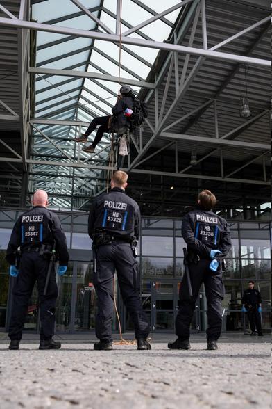 Eine Person seilt sich ab von einer Konstruktion aus Metall, die ein Glasdach hält. Davor stehen drei Polizist:innen und sehen nach oben zu der Person. Im Hintergrund ist die Glasfassade des Gebäudes zu sehen.
Bild: Pay Numrich