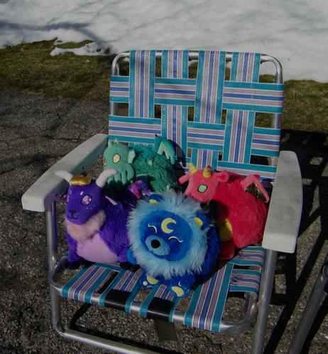 Four plushies sitting on a blue, lavender and teal lawn chair. One plushie is a purple sheep/goat (Cosmic Demon), another is a blue lion with a moon symbol and closed eyes, and behind them is a green Cthulhu and a red dragon plush, who is hiding behind the other plushies.