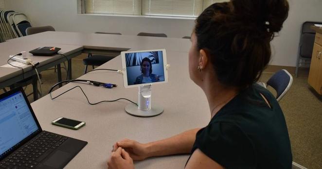 photo of a woman sitting at a table with her laptop, speaking via teleconference to a woman on an ipad screen