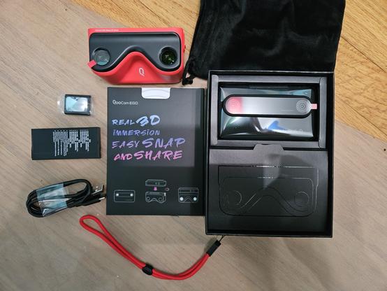 A disassembled view of the packaging for the Kandao QooCam Ego. The camera is still in the box with the lenses facing upwards. Outside of the box is the insert, the cloth holding bag, the 3D viewer for the camera, a small selfie mirror, the battery, a USB C cable, and a wrist strap.