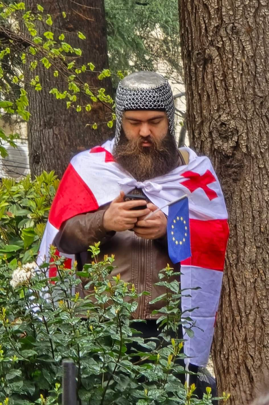 Georgian protestor wearing traditional armor and using Georgian flag as cloak. He has prominent beard similar to likely medieval knights had. He holds small EU flag in his hand and looks into his smartphone