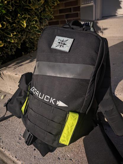 Black backpack with a grey logo that reads Go Ruck. There are two yellow reflector straps on the bag.