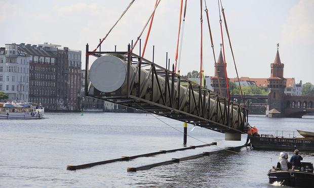 Wikipedia-Foto aufgenommen von Ralf Steeg: Ein sehr großes zu allen Seiten verschlossenes Glasfaserrohr hängt an Kranseilen und wird in die Spree gelassen. Im Hintergrund ist die Oberbaumbrücke zu sehen.