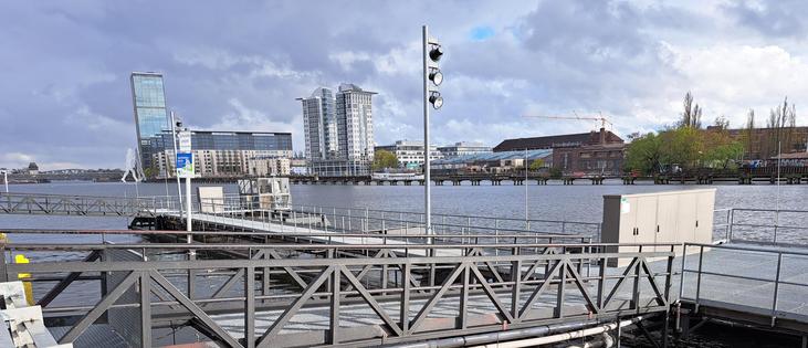 Vom Regenwasserrückhaltespeicher ist  über der Wasseroberfläche nicht mehr zu sehen, als eine im Wasser schwimmende, metallgefertigte Steganlage mit Geländer. Im Hintergrund sind auf der anderen Spreeuferseite die Treptowers zu sehen.