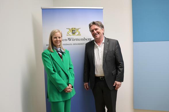 Das Foto zeigt zwei Personen, die vor einem Roll-Up-Banner stehen. Auf dem Banner ist das Logo des Sozialministeriums zu sehen. Rechts im Bild zu sehen ist Minister Manne Lucha, links daneben steht die neue Chefin der Regionaldirektion Baden-Württemberg der Bundesagentur für Arbeit, Martina Musati. Beide lachen und schauen in die Kamera.