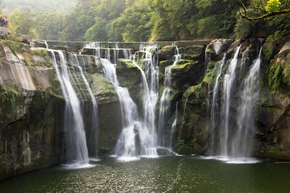The Shifen waterfall is a wide waterfall cascading over rocky cliffs into a serene pond, surrounded by lush greenery with spectators on an observation deck to the left.