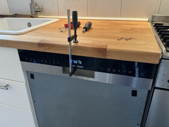 A kitchen scenario with a wooden butcher block countertop being secured by a bar clamp above a Siemens dishwasher, next to a sink and gas stove. Tools and screws lie on the countertop.