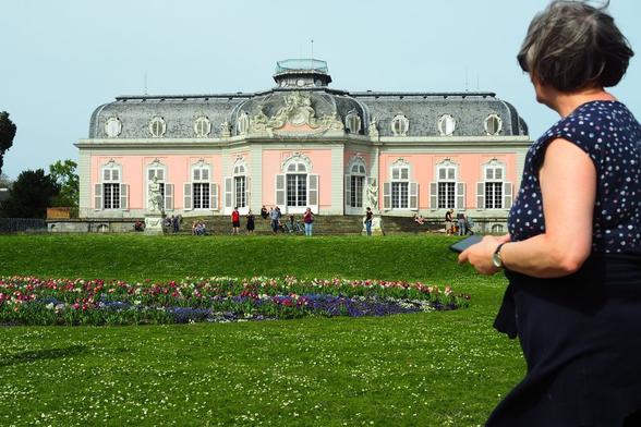Barockes Schloss mit rosa Fassade auf einer Anhöhe in einem Park mit blühenden Frühlingsblumen. Davor halb von hinten Betrachterin. Gesättigte Farben, azurfarbener Himmel.