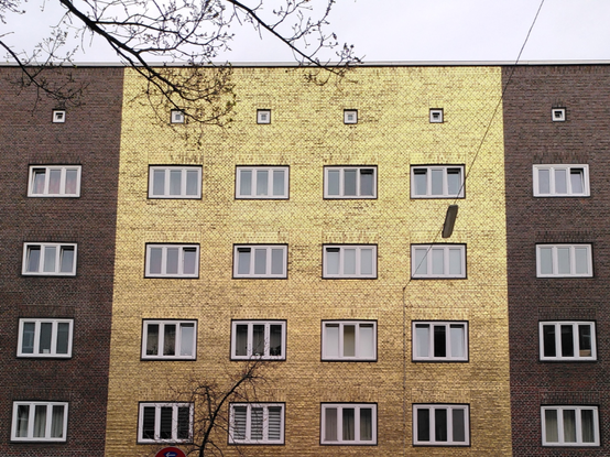 You are looking at the plain front wall of three connected multistorey flats with five rows of windows. The wall is built from bricks: The left and right units are built with red bricks but the middle unit stands out because it is built with yellow bricks which are covered with golden paint — quite shiny in the golden morning sun.