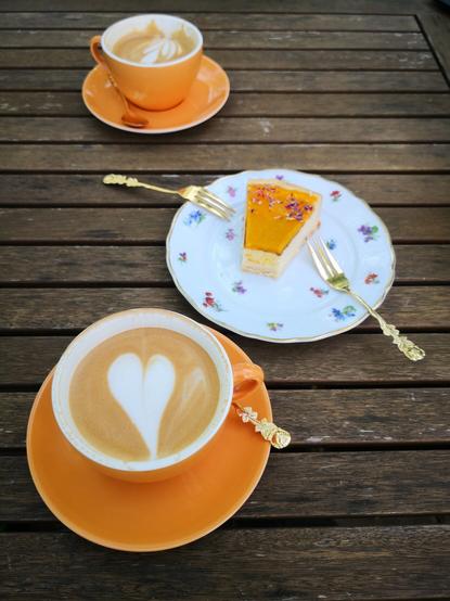 Zwei große Cappuchino in orange-ocker Tassen mit weißem Inneren, einander gegenüberstehend auf einem hölzernen Gartentisch, dazwischen ein Kuchenstück mit zwei Kuchengabeln auf einem Tellerchen in altem Blümchendekor und Goldkantemit.

Die Cappuchino haben eine feine Crema, im Milchschaum der vorderen Tasse ein Herz, das inzwischen durch das Trinken länglich verzogen ist. Gleiches gilt für die andere Tasse mit ihrem Farn-Motiv.

Das inzwischen angebrochene Kuchenstück ist ein Käsekuchen mit Mangodecke.