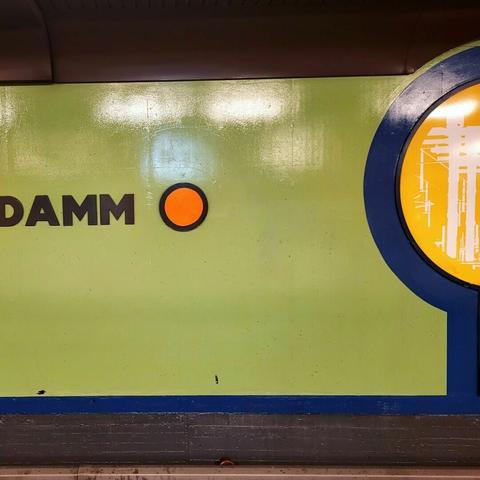 Grasgrüne Wand mit Grafiksymbolik. Links im Bild angeschnitten sind Lettern eines unvollständigen Worts "Damm", rechts davon eine rote kreisrunde Farbfläche. Das rechte Bildrand ist eine hellgelbe große Kreishälfte umgeben mit einer dicken dunkelblauen Kontur. Am unteren Bildrand ist ein Bahngleisbett erkennbar