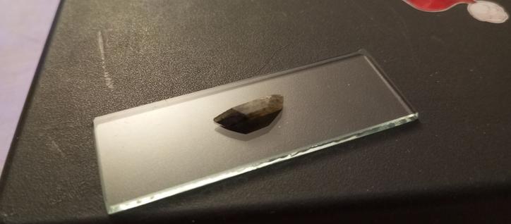 Фото камня на предметном стекле. Он огранён в форме, напоминающей трапецию, с широкими основаниями и небольшой высоты и толщины. По большей части он желтоватый, но один край - более тёмный, почти чёрный. Камень немного прозрачный, видно, что в нём есть слои.