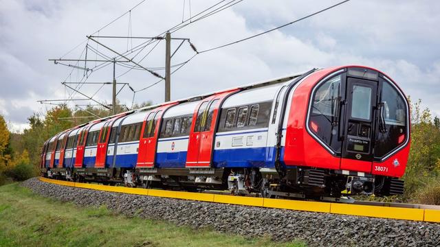 The new Piccadilly rolling stock of the London underground hurtles around a corner on a test run. It is in a rather bushy, grassy area, with the test track a bold yellow on an island of grey pebbles.