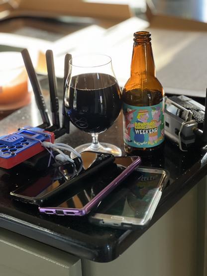 A 12 ounce brown bottle of beer with pastel colored people and lettering. three smartphones and two wardrving wigs surround it and a tulip glass.