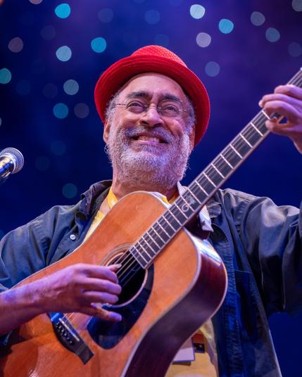 Vance Gilbert plays his guitar while looking at the audience behind the camera. He is smiling broadly while wearing a bright red hat.