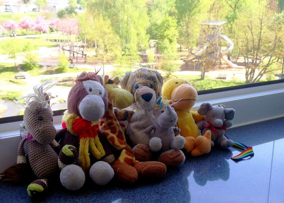 Die Plüschies Strickpony Zuppy, Reisepony, Giraffe kami, Tiger Hobbes mit Einhorn Carola auf dem Schoss, Ente Pöttke und Elefanty Elfi sitzen aneinander gereiht auf der Fensterbank des geöffneten Fensters. Im Hintergrund die Aussicht aus dem Büro, vor lauter Sonne überblendet: der Erba-Spielplatz, rosa blühende Bäume, …