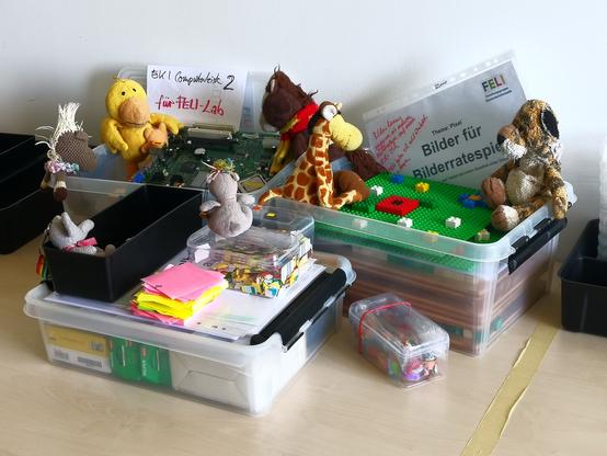 Die Plüschies untersuchen Kisten mit informatischem Erfahrungsmaterial: zu sehen sind u.A. ein Motherboard, eine Steckplatte mit quadratischen Klemmbausteinen, Zettelschnippsel, Spielfiguren