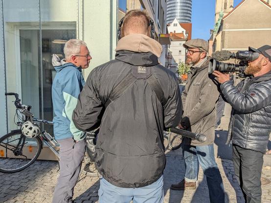 Der MDR interviewt Michael vom Radentscheid Jena
Außer Michael sind 3 Personen des MDR zu sehen: der Journalist und je ein Mitarbeiter für Kamera und Ton.
Die Szene wurde am Ostende der Saalstraße in Jena fotografiert; Blickrichtung nach Westen. Vor einem der abgebildeten Häuser sieht man ein abgestelltes Fahrrad, an dessen Lenker ein Fahrradhelm hängt.
Das Interview wurde bei sonnigem Wetter und blauem Himmel geführt (beides auf dem Foto zu erkennen).