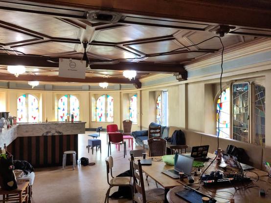 Une grande salle lumineuse avec des vitraux dans le fond, quelques tables et chaises avec des ordinateurs à l'avant plan, un bar sur la gauche. Accrochée sur une rallonge électrique suspendue, une affiche mentionnant l'AG de Neutrinet.